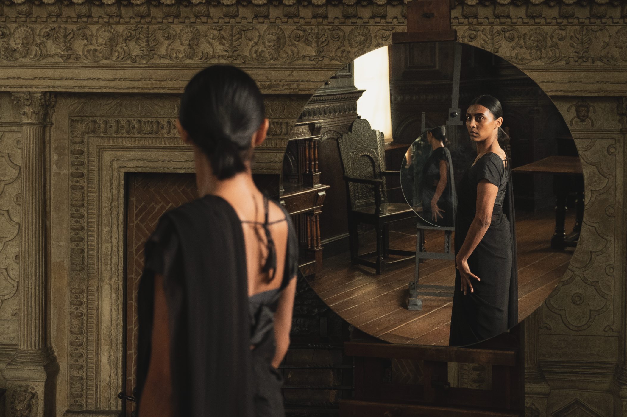 Lumen - Woman in black dress looking at herself in a large circular mirror in an ornate historic interior